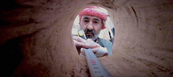 The camera looks up at a man with a short salt and pepper beard wearing a red headscarf and measures the inside of a hollow wooden log, with the opening framing his face.